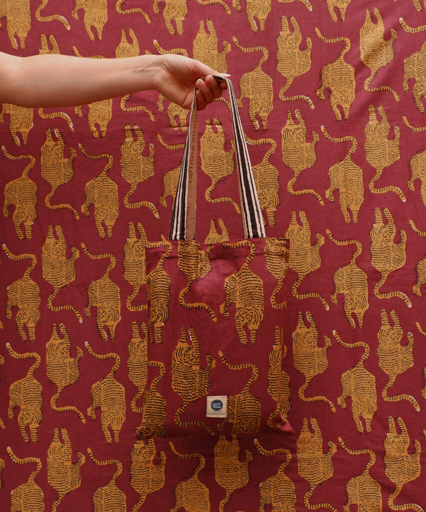 Borsa in tessuto con stampa di tigri gialle su sfondo (tovaglia) rosso, perfettamente mimetizzata con lo sfondo identico; una mano tiene la borsa per i manici a righe.