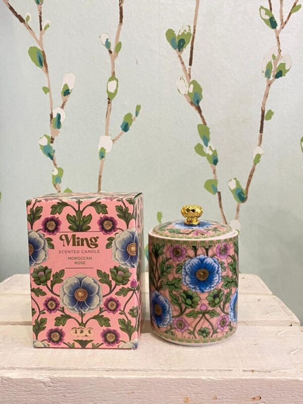 Candela profumata 'Ming' alla rosa in contenitore decorato con motivi floreali blu e rosa, accanto alla confezione coordinata su uno sfondo con parete a rami stilizzati.