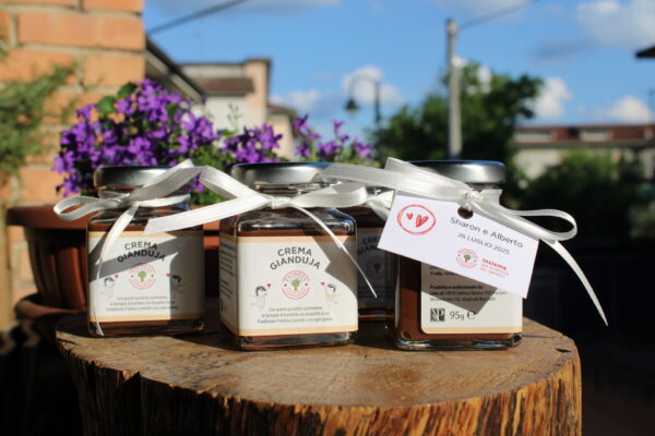 Vasetti di crema gianduja da 95g con etichetta personalizzata per matrimonio, decorati con fiocchi bianchi e disposti su un tagliere in tronco di legno. Aall'aperto, con fiori viola sullo sfondo.