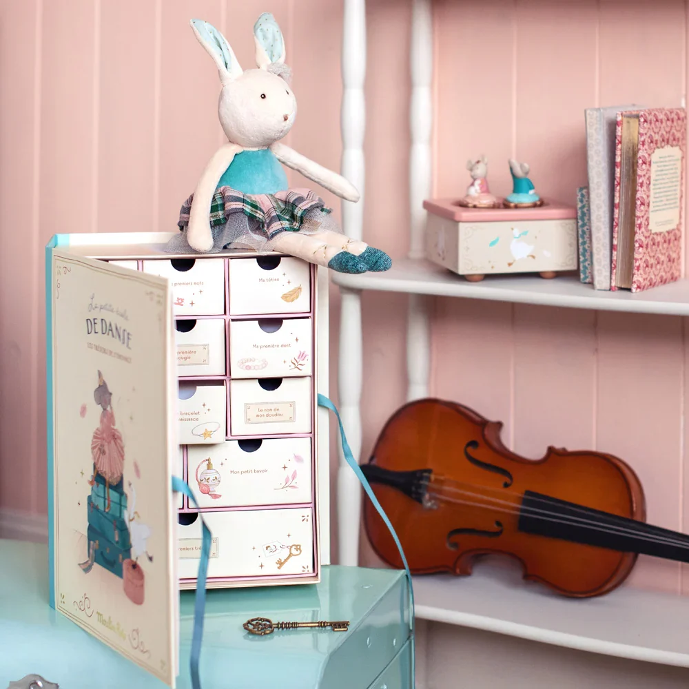 Scatola ricordo Moulin Roty con cassettini illustrati e coniglietta in tutù seduta sopra, ambientata in una cameretta rosa con violino e libri