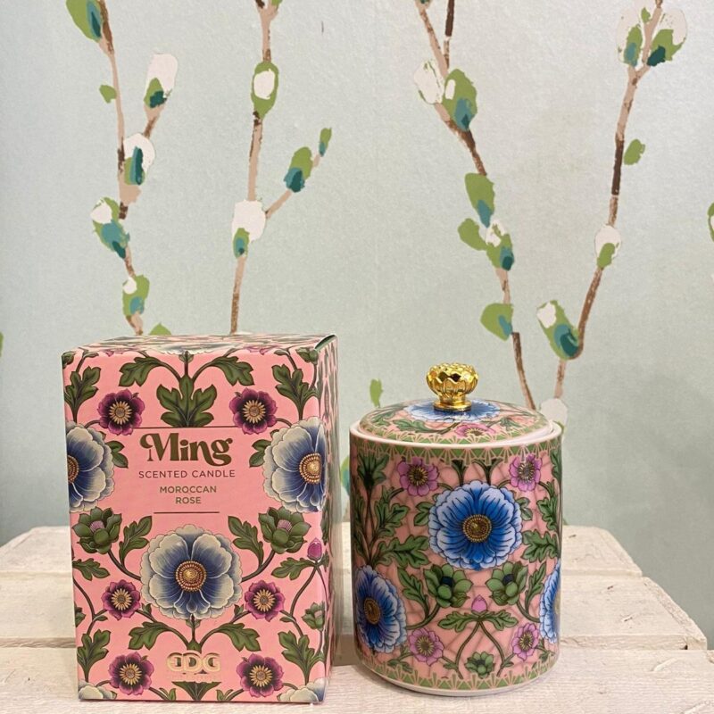 Candela profumata 'Ming' alla rosa in contenitore decorato con motivi floreali blu e rosa, accanto alla confezione coordinata su uno sfondo con parete a rami stilizzati.